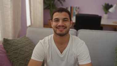 Genç İspanyol adam, evinde gülümseyerek elleriyle kalp hareketi yapıyor.