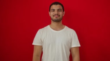 Beyaz tişörtlü, kendine güvenen, pozitif gülümseyen mutlu ve rahat bir şekilde izole edilmiş kırmızı arka planda duran İspanyol genç adam.