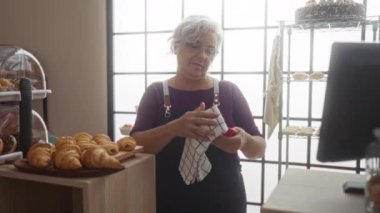 Orta yaşlı, gri saçlı bir kadın, sıcak bir fırında tabak yıkıyor, hamur işleri sergileniyor, kapalı gişenin etrafında kuruluyor.