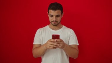 Adam gülümsüyor ve işaret veriyor parmaklarıyla akıllı telefon kullanıyor - genç İspanyol adam kırmızı arka planda izole edilmiş