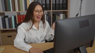 Woman smiling during videoconference at office desk with bookshelves in background, wearing eyeglasses and white blouse, brunette female in workplace environment.