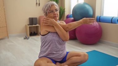 Yetişkin bir kadın spor salonundaki yoga minderinde egzersiz topları ve arka planda spor aletleriyle esneme egzersizleri yapıyor.