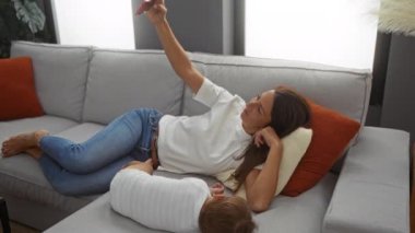 Küçük oğluyla rahat bir oturma odasında kanepede uzanan ve selfie çeken bir kadın. Dairedeki modern koltukta, yastıklarla birlikte eğlenen bir kadın..