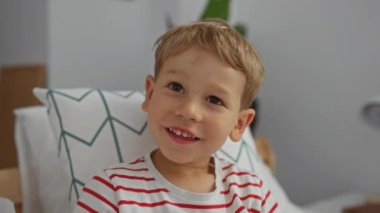 Çizgili tişörtlü sarışın çocuk rahat bir yatak odasında bir yatağa oturur, gülümser ve içeride oynar, sevimli çocukluk anları sergiler..