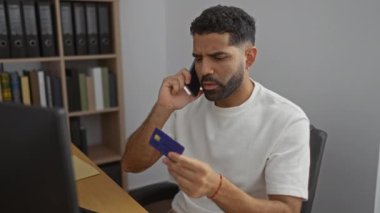 Ofisteki genç adam elinde kredi kartıyla endişeli görünüyor.