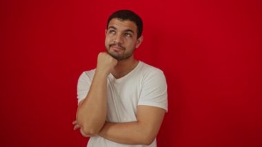 Beyaz tişört giyen genç İspanyol adam ciddi bir yüz ve çene üzerinde düşünme ve düşünceli, bir fikir ya da soru izole kırmızı arka plan üzerinde
