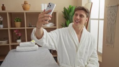 Genç adam spa sağlık merkezinde selfie çekerken beyaz bornoz giyerken rahat bir şekilde masaj masasında bitki ve raflarla oturuyor.