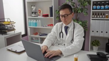 Beyaz önlüklü Asyalı erkek doktor, tıbbi ekipmanlar ve dizüstü bilgisayarla çevrili bir hastane kliniğinde masasında otururken gözlüklerini çıkarıyor ve gözlerini ovuyor..