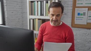 Ofisteki İspanyol adam modern işyerindeki bilgisayarda doküman okuyor, raflar ve grafiklerle çevrili, konsantrasyon ve neşe ifade ediyor..