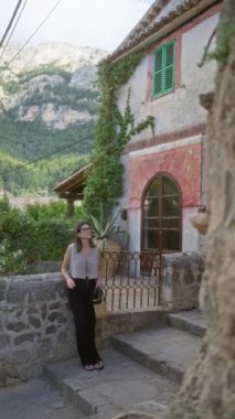 Deia, Mallorca 'da dışarıda duran genç bir kadın, doğayla çevrili yeşil kepenkleri olan büyüleyici taş bir evin yanında duruyor ve elinde kamış bir çanta tutan, güzel bir dağ zemininde.