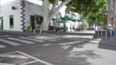 Lanzarote 'daki bulanık manzara, bankta oturan bir adamla İspanya ve beyaz binanın yanındaki yeşil şemsiyelerin altında yemek yiyen insanlar.