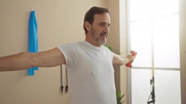 Olgun bir adam sağlık merkezinde halterlerle evde egzersiz yapıyor, dingin bir ortamda zindelik ve sağlık gösteriyor..