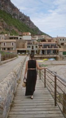 Port de valldemossa, mallorca, İspanya 'da ahşap geçitte yürüyen bir kadın, elinde bir hasır çanta tutuyor, kırsal evler ve bulutlu bir gökyüzü altında dağlık araziyle çevrili.