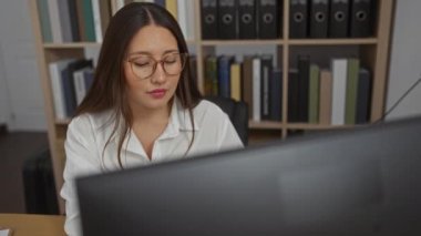 Ofis ortamında çalışan İspanyol bir kadın, bilgisayar üzerine odaklanmış, ciltler ve kitaplarla dolu bir kitap arkaplanı var.