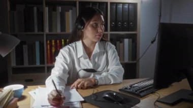 Ofisteki bilgisayarda çalışan, not alan ve kulaklık takan genç bir kadın, arka planda klasörler ve kitaplarla dolu bir ofis ortamında.