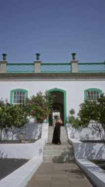 Otel terasında duran bir kadın açık mavi gökyüzü, yeşil bitkiler ve arka planda yeşil çizgileri olan beyaz bir bina..