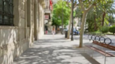 Ağaçlar ve banklarla kaplı güneşli bir kaldırım boyunca yürüyen odaksız insanların olduğu açık hava sahnesi.