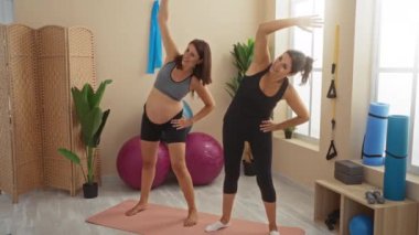 Hamile kadın iyi donanımlı bir spor salonunda kadın antrenörle yoga yapıyor.