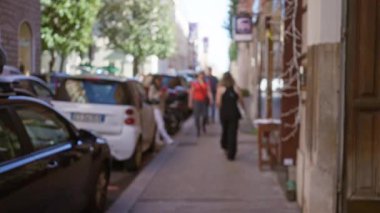 Roma 'nın güneşli bir caddesinde yürüyen insanlar bulanık hareketlerle ve ağaçların altına park edilmiş arabalarla bir bokeh etkisi yaratıyorlar.