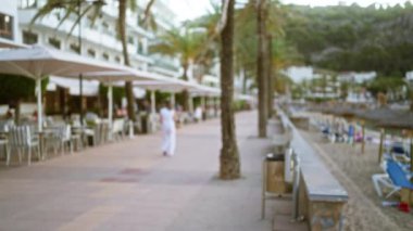 Gün batımında, Mallorca, İspanya 'da sahil boyunca uzanan açık kafeler ve palmiye ağaçları ile bulanık yürüyüş yolu. Huzurlu sahil kenarında yürüyen beyaz giysili yalnız bir kadın..