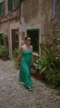 Yeşil elbiseli ve güneş gözlüklü bir kadın Valldemossa, Mallorca 'da büyüleyici bir kaldırım taşı caddesi boyunca yürüyor.