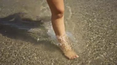 Kumsalda sığ sularda yürüyen bir kadın, gün ışığı altında bacaklarını ve ayaklarını gösteren temiz kumlarla..