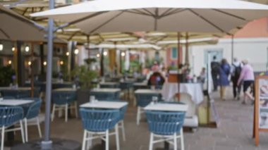 Villefranche-sur-mer Fransa 'da mavi sandalyeli ve şemsiyeli açık hava kafe aydınlık bir sokakta yürüyen ve eğlenen bulanık insanları gösteriyor.