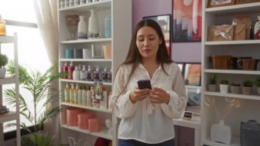 Ev dekorasyonu mağazasında genç bir kadın şık iç dekorasyonlarla çevrili telefonunda geziniyor..