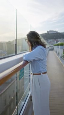 Kadın, güneşli bir günün tadını çıkarıyor. Gözlük takıyor ve şık giyiniyor. Arka planda tarihi bir manzara ve cam parmaklıklar var..