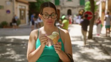 Güneşli Valldemossa, Mallorca, İspanya 'da dondurmadan hoşlanan bir kadın, gözlük takıyor ve arka planda insanların olduğu hareketli bir sokak sahnesiyle çevrili yeşil bir elbise giyiyor.