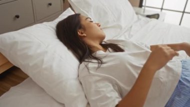 Yatak odasında beyaz yastıklar ve çarşaflarla uzanan, günlük beyaz tişört ve mavi şort giyen, evinde huzur içinde dinlenen genç bir kadın.