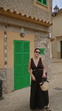 İspanyol asıllı genç bir kadın Mallorca İspanya 'nın limanında güneş gözlüğü takıyor ve yeşil panjurlu bir binanın yanında elinde kamış çantası tutan siyah bir elbise giyiyor.