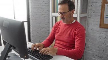 Ofiste bilgisayar başında çalışan yaşlı bir adam, modern iş yerindeki kitaplıklarla birlikte masaüstünde otururken omuz ağrısı çekiyor..