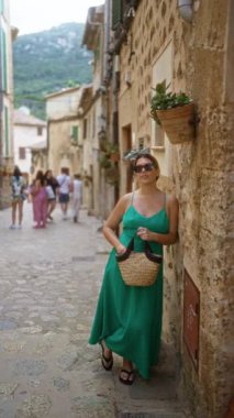 Yeşil elbiseli genç İspanyol kadın Valldemossa, Mallorca 'nın pitoresk caddesinde taş bir duvara yaslanıyor. Arka planda dağlarla birlikte İspanya' da..