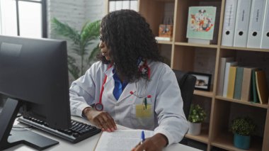 Kıvırcık saçlı Afro-Amerikalı genç bir kadın doktor klinikteki bir bilgisayarda çalışıyor. Etrafı kitaplık ve tıbbi dosyalarla çevrili..