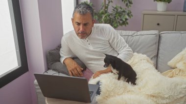 Orta yaşlı İspanyol bir adam oturma odasında dizüstü bilgisayar ve chihuahua köpeğiyle dinleniyor..