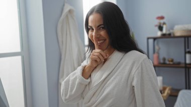 Sessiz bir jest yapan genç bir kadın, arka planda sağIıklı eşyalar bulunan rafların bulunduğu sakin bir kaplıca odasında bornoz giyerken parmağını dudaklarına koyuyor.