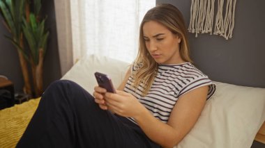 Genç, beyaz bir kadın yatak odasında, bir yatakta oturuyor, akıllı telefonunu evde kullanıyor..