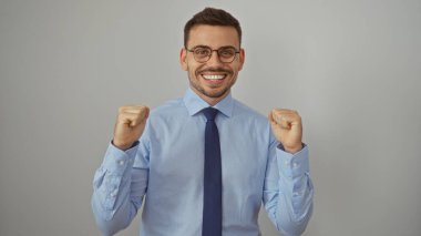 Young hispanic man with a beard in a blue shirt and tie, smiling confidently with fists raised, standing isolated against a white background. clipart