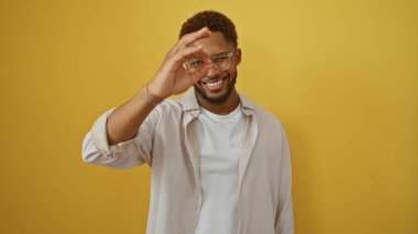 Genç adam gözlerinin önünde işaret veriyor, beyaz gömlek giyiyor, izole edilmiş sarı bir arka plana karşı duruyor ve güvenle gülümsüyor.