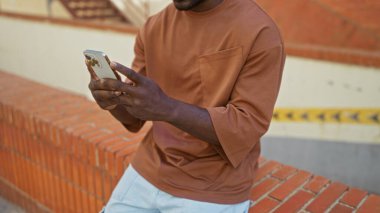 Şehir sokaklarında akıllı telefon kullanan yakışıklı Afrikalı bir adam tuğla duvarda oturuyor.