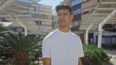 Beyaz tişörtlü yakışıklı bir İspanyol, güneşli bir günde yeşil bitkiler ve modern şehir mimarisi ile çevrili canlı bir şehir parkında dikiliyor..