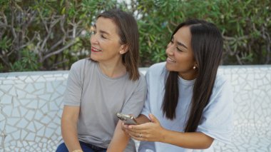 Anne ve kızı açık bir bankta birlikte gülümsüyor.