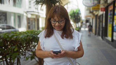 Olgun bir İspanyol kadın şehir merkezinde telefonuna gülümseyip açık havada bir bağlantı ve teknoloji anını yakalıyor..