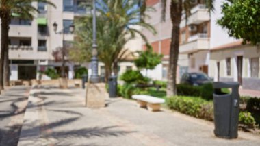 Palmiye ağaçları, banklar ve arka planda çözünmüş binalar ve yeşillik olan bir çöp tenekesinin yer aldığı bulanık şehir manzarası bir bokeh etkisi yaratıyor