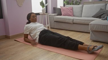 Güzel, Afro-Amerikan bir kadın. Kıvırcık saçlı. Evinin rahat oturma odasında yoga minderinde egzersiz yapıyor..