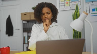 Kıvırcık saçlı, modern ofis ortamında dizüstü bilgisayar üzerinde çalışırken derin düşünen Afro-Amerikalı genç bir kadın..