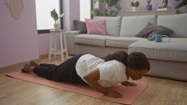 Güzel, Afro-Amerikan bir kadın oturma odasında şınav çekiyor. Parlak bir dekor ve pencereden içeri ışık giriyor..