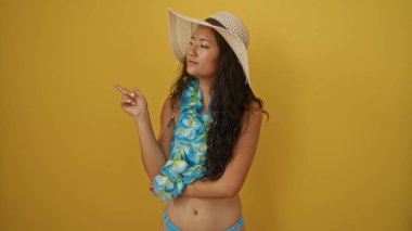Güzel, genç Asyalı bir kadın sarı bir arka planı işaret ediyor. Şapkalı ve Hawaii zambağı takıyor.