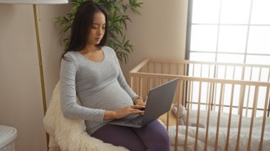 Hamile bir kadın rahat bir yatak odasında dizüstü bilgisayar kullanıyor, beşikte oturuyor, sandalyede oturuyor, günlük giysiler giyiyor ve evde rahat bir şekilde görünüyor..